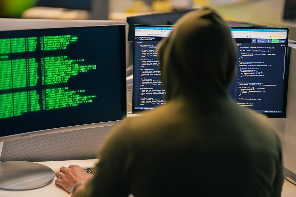 Hacker in hoodie working on multiple computer screens