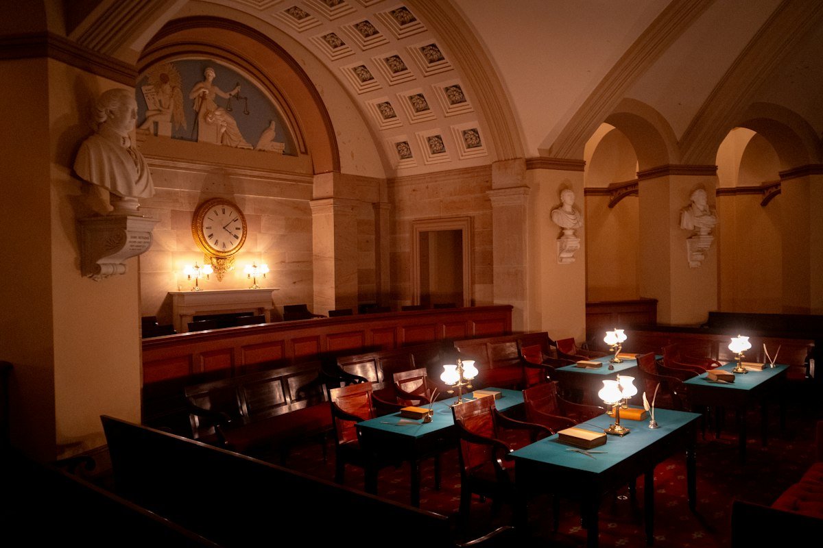 A historic courtroom with wooden benches, desks with lamps, and marble busts