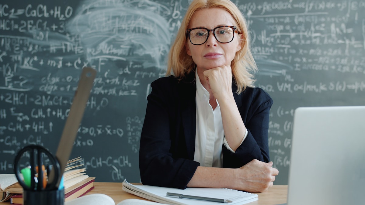 Teacher sitting at a desk with chalkboard formulas